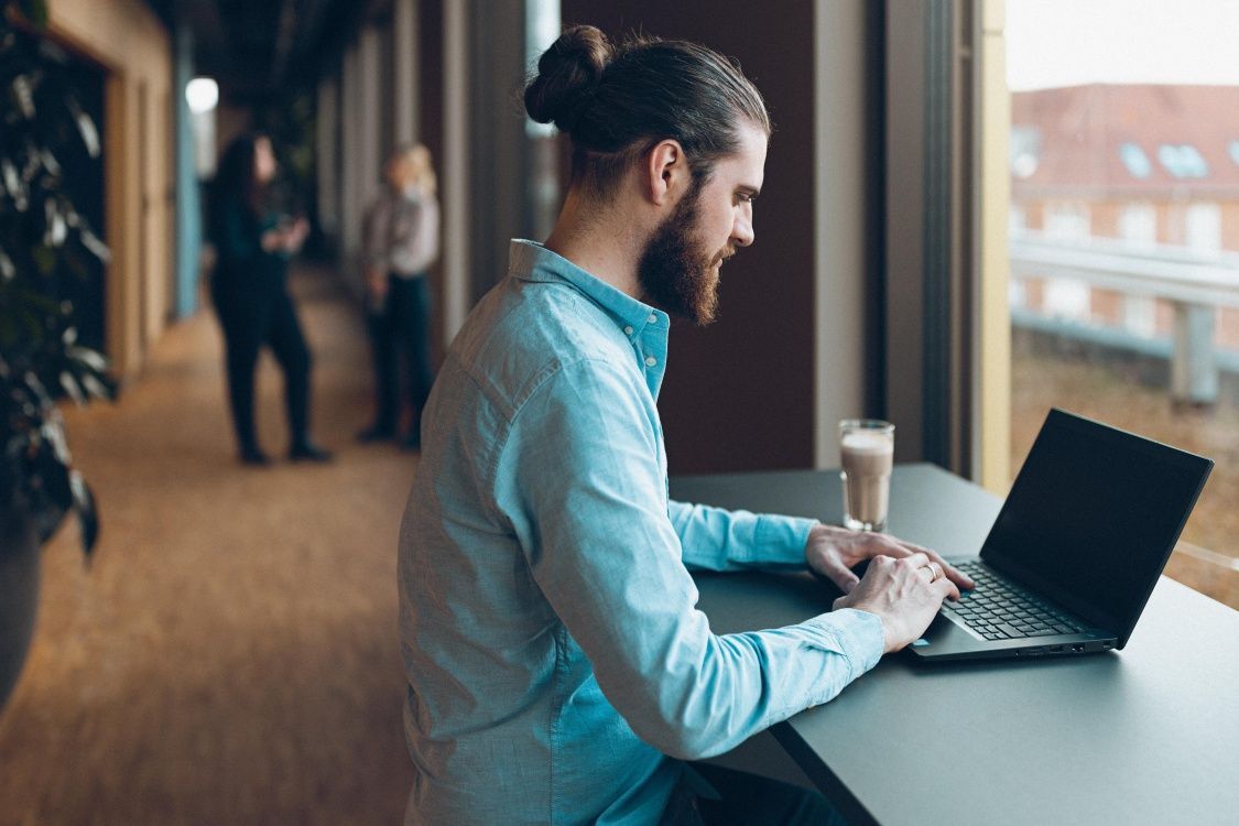 Vīrietis ar matu sakārtojumu copē sēž pie galda modernā birojā un strādā ar portatīvo datoru. Fonā redzami kolēģi.