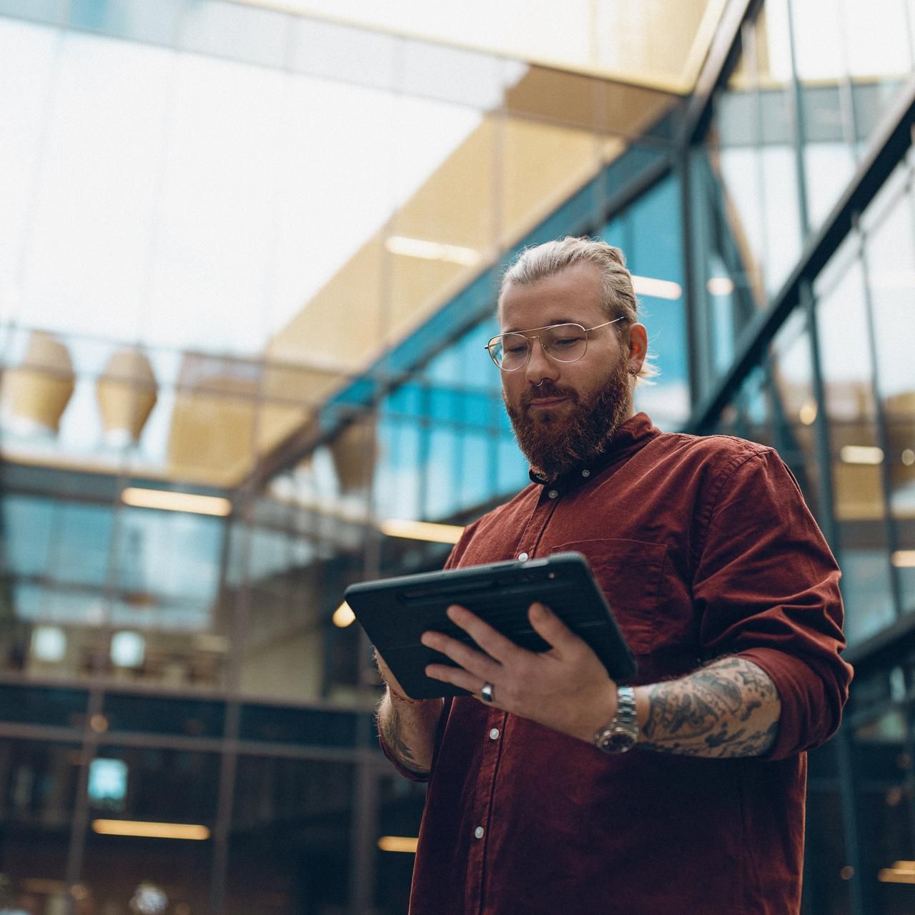 īrietis ar brillēm un bārdu strādā ar planšetdatoru uzņēmuma dārza telpā biroja ēkas centrā.