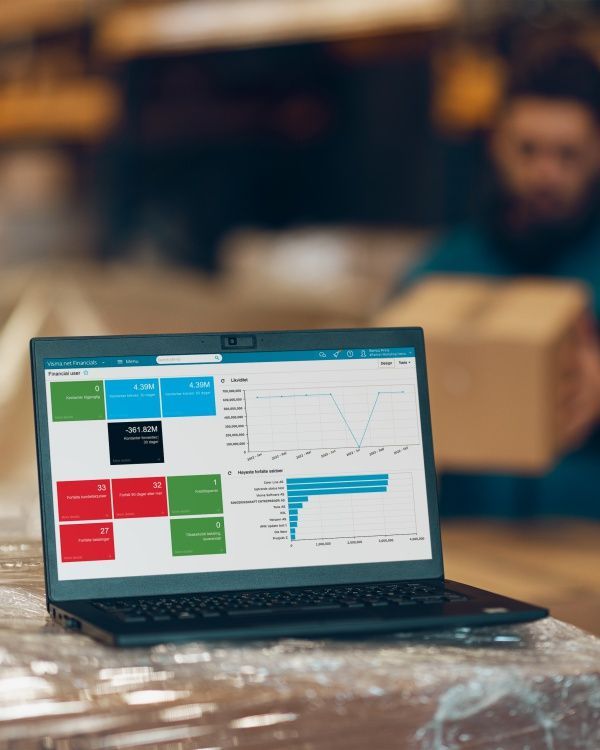 Portatīvais dators uz plēvē ietītas paletes noliktavas fonā. Ekrānā redzami finanšu grafiki un datu paneļi.