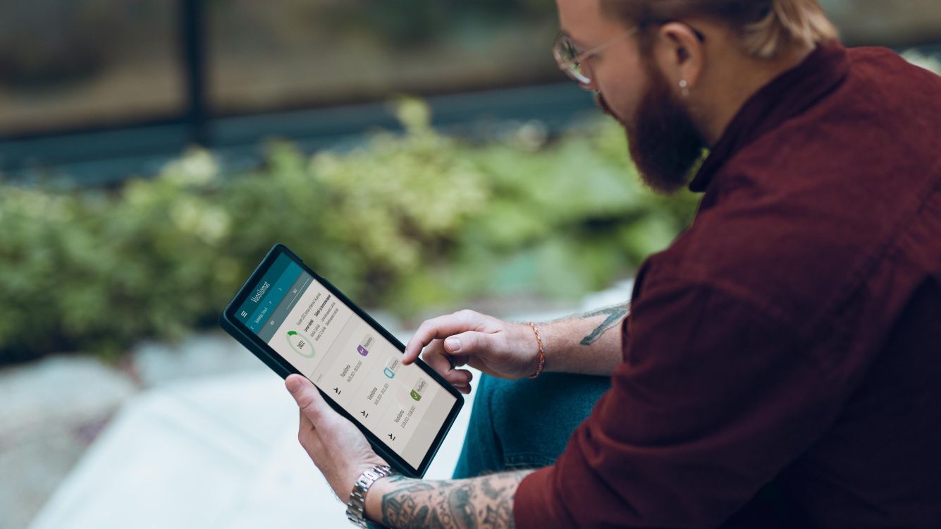 Vīrietis ar tetovējumiem uz rokām brīvā dabā lieto planšetdatoru, kurā redzama personāla vadības sistēmas atvaļinājumu sadaļa.