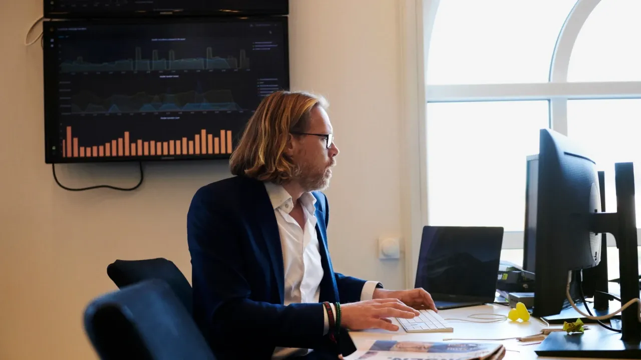 Vīrietis birojā strādā pie datora, fonā redzami datu grafiki uz ekrāna.