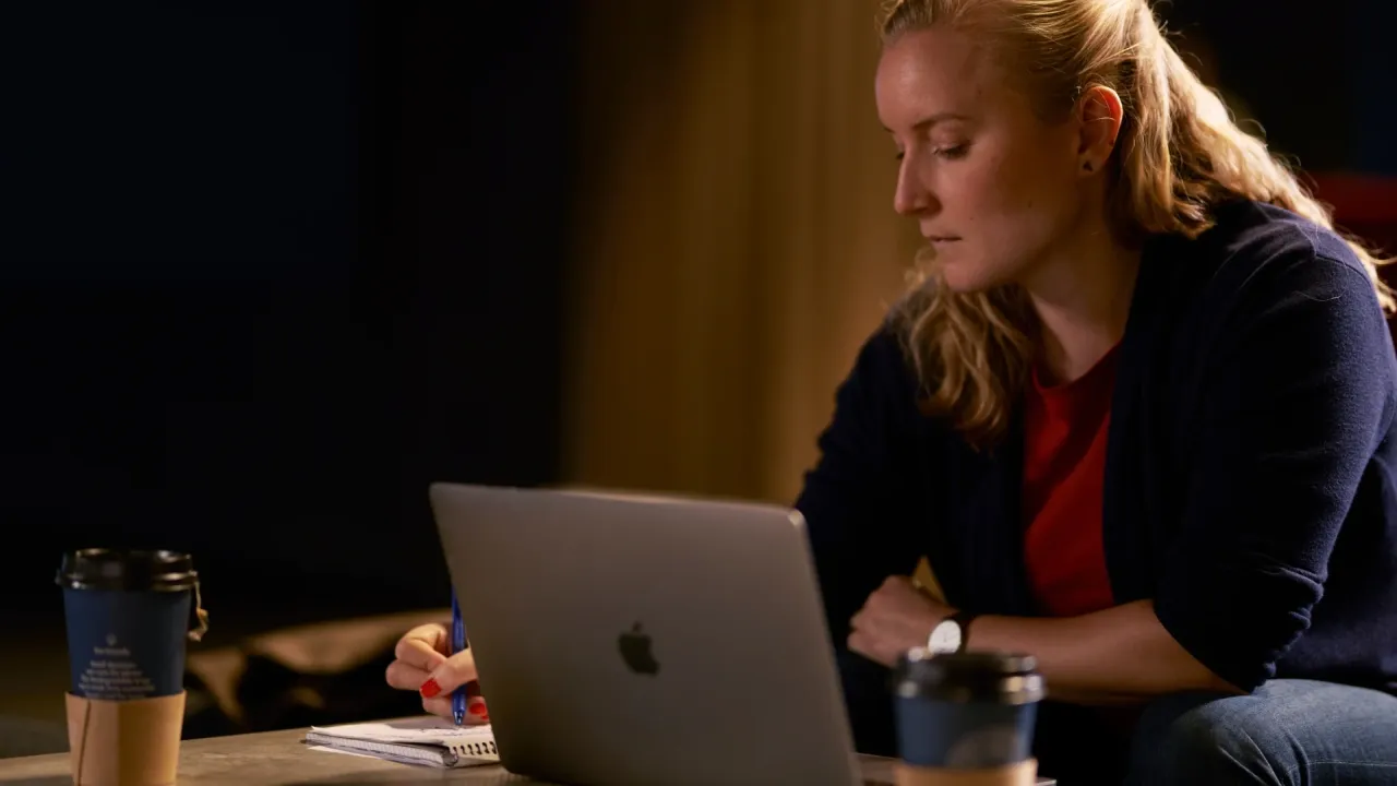 Sieviete strādā pie klēpjdatora un veic piezīmes kladē.