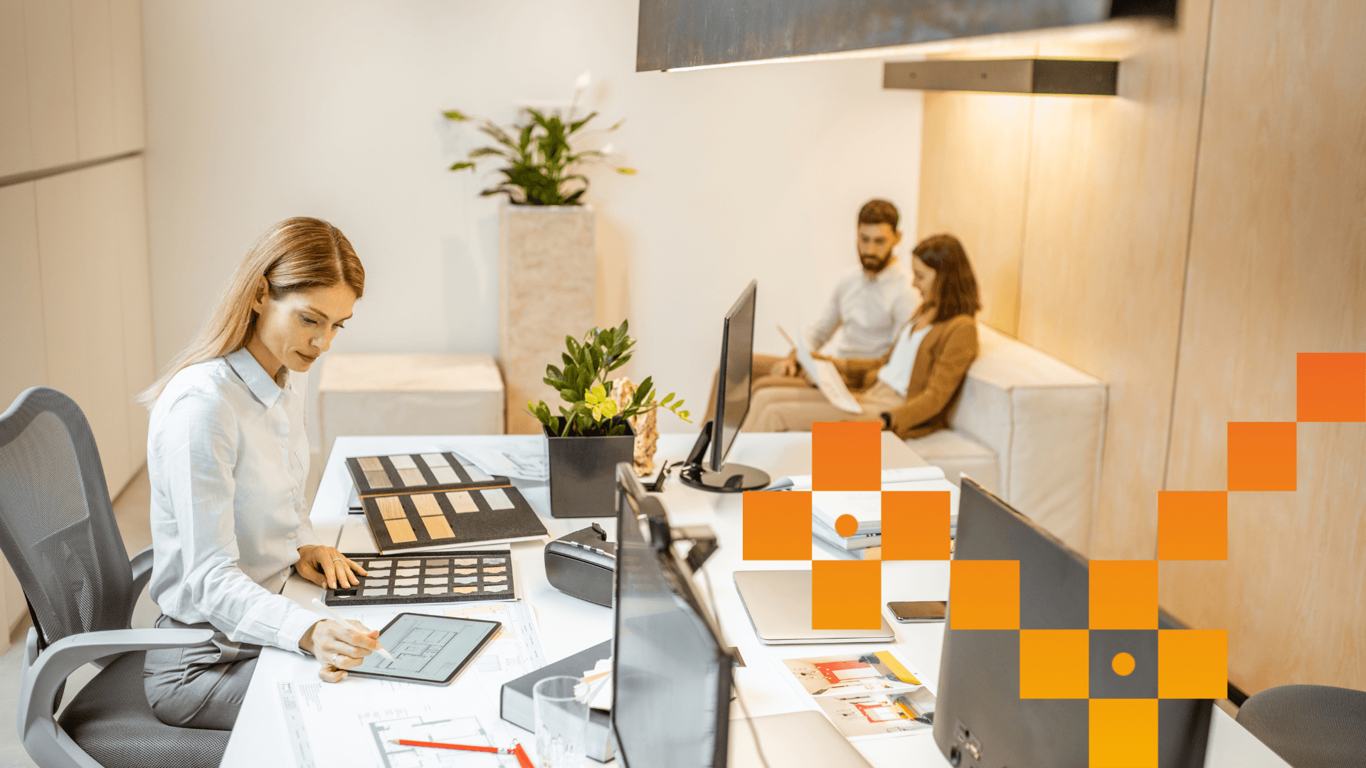 Sieviete birojā strādā pie galda ar plānotāju un planšetdatoru, fonā uz dīvāna sarunājas divi kolēģi. Moderns interjers ar gaišām mēbelēm un telpaugiem.