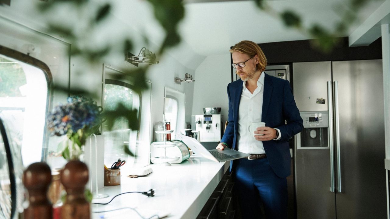 Vīrietis elegantā tērpā strādā pie klēpjdatora virtuvē, tur rokā kafijas krūzi.