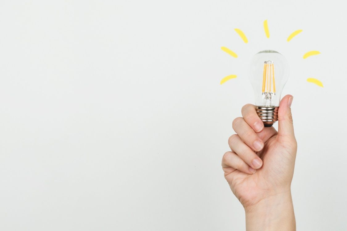 Roka tur caurspīdīgu kvēlspuldzi uz balta fona, virs kuras uzzīmēti dzelteni gaismas stari.