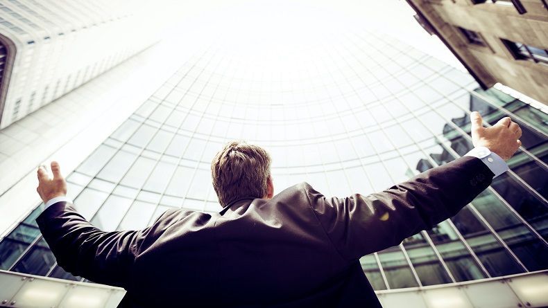 Skats no lejas uz vīrieti uzvalkā, kurš stāv ar plaši atplestām rokām pretī augstam, stiklotam debesskrāpim un saules gaismai.