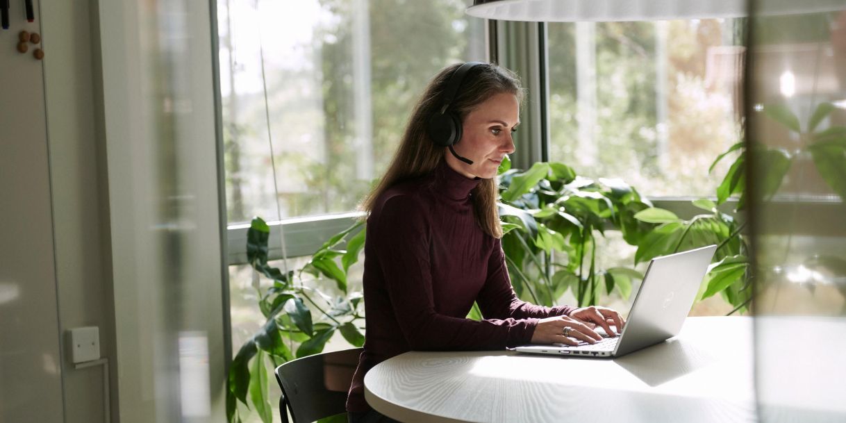 lady-with-laptop.jpg Sieviete ar austiņām un mikrofonu strādā pie klēpjdatora gaišā, zaļumos apjoztā biroja telpā.