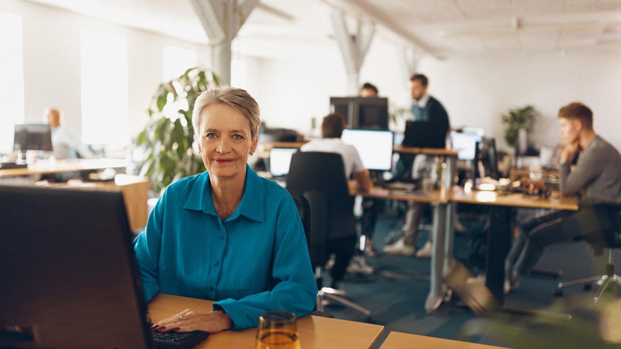 Pieredzējusi sieviete sēž pie galda birojā un skatās tieši kamerā, fonā strādā citi darbinieki.
