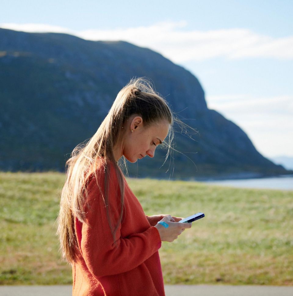 Sieviete lauku ainavā skatās uz viedtālruni, izmantojot Jumis My Assistant risinājumu.