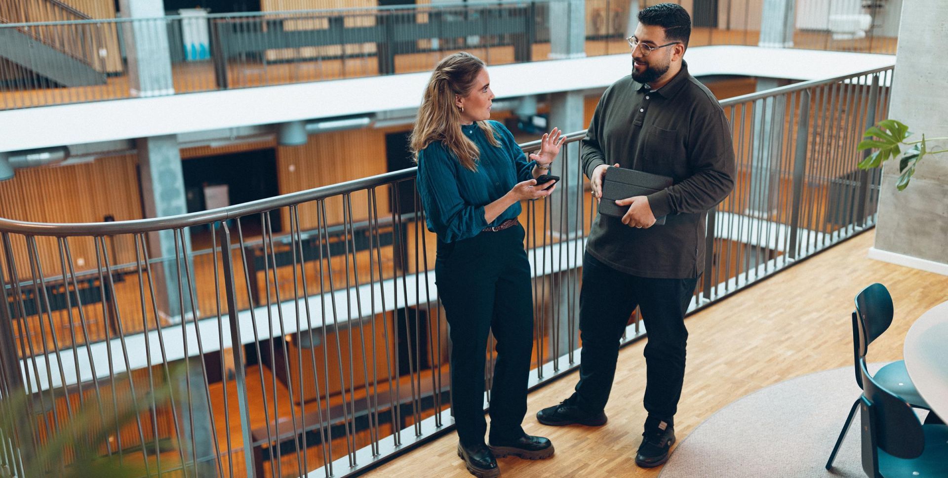 Divi kolēģi sarunājas modernā biroja ēkas gaitenī, apspriežot darba jautājumus.